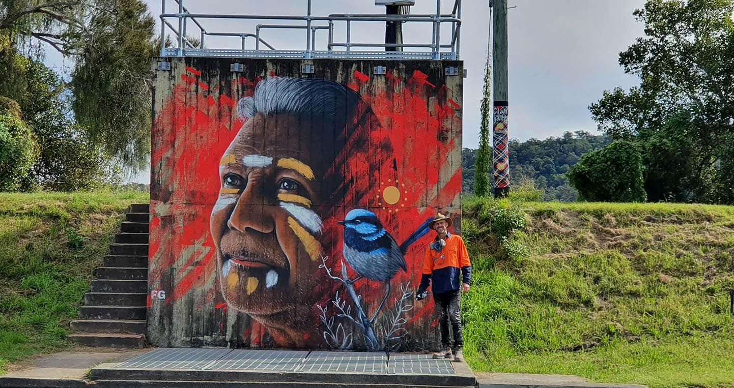 Street artist Austin with the completed mural depicting Yaegl Elder Uncle Ron Heron. The mural can be seen on the old pump station in River Street, Maclean. Image: Lynne Mowbray